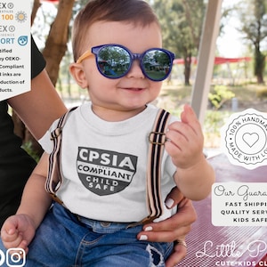 K&ouml;nnte beinhalten: Ein l&auml;chelndes Baby mit blauer Sonnenbrille und einem wei&szlig;en T-Shirt mit dem Text "CPSIA COMPLIANT CHILD SAFE". Das Baby tr&auml;gt auch Hosentr&auml;ger. Das Bild enth&auml;lt Text &uuml;ber die OEKO-TEX-Zertifizierung und handwerkliche Qualit&auml;t.