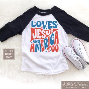 May include: A white and black raglan style t-shirt with the words "LOVES JESUS AND AMERICA TOO" in red and blue. The shirt is paired with white sneakers.