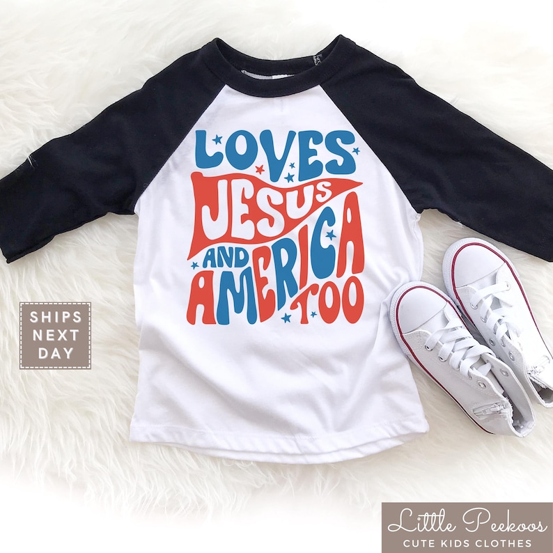 May include: A white and black raglan style t-shirt with the words "LOVES JESUS AND AMERICA TOO" in red and blue. The shirt is paired with white sneakers.