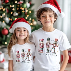 May include: Two children wearing white t-shirts with a Christmas nutcracker design. The shirts say "Tis the Season" and feature a nutcracker holding a sign that says "Help!"