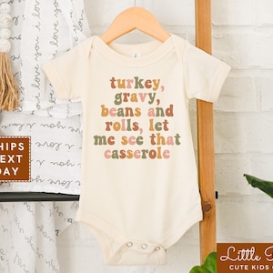 May include: A beige baby onesie with the text "turkey, gravy, beans and rolls, let me see that casserole" in a retro font with brown, pink, and green colors.