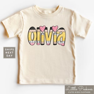 May include: A cream colored t-shirt with the name "Olivia" spelled out in yellow, black, and pink pencil designs. Two pink hearts are above the name.