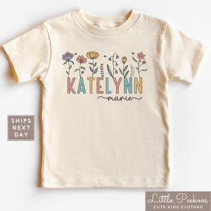 May include: Cream-colored t-shirt featuring the name "KATELYNN" in colorful block letters, with "marie" in cursive below. A floral design with various flowers and leaves is above the name. The shirt is from "Little Peekoos Cute Kids Clothes."
