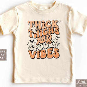 May include: Cream-colored t-shirt with the text "Thick Thighs and Spooky Vibes" in orange and white. The design includes a ghost, bats, and a jack-o'-lantern, perfect for Halloween. The shirt is a children's clothing item.