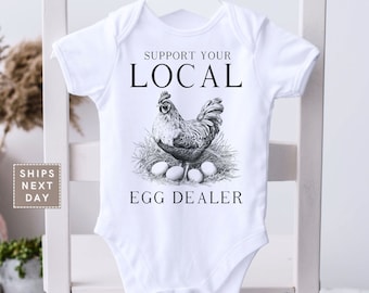May include: White baby onesie with a black and white illustration of a rooster and eggs. The text "Support Your Local Egg Dealer" is printed on the onesie.