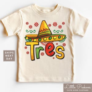May include: A cream-colored t-shirt featuring a sombrero design with the word "Tres" in yellow, green, and red. The sombrero is yellow with green and red stripes and green and yellow pom-poms. Red flower graphics are scattered around the sombrero.