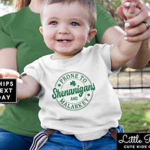 May include: White baby onesie with a green circle graphic that says "Prone to Shenanigans and Malarkey".