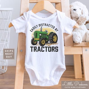 K&ouml;nnte beinhalten: Wei&szlig;er Baby-Body mit einer gr&uuml;nen Traktor-Grafik und dem Text "EASILY DISTRACTED BY TRACTORS". Der Traktor hat gelbe R&auml;der und ist vor einem gelb-orangen Hintergrund platziert. Der Body h&auml;ngt an einem Holzb&uuml;gel.