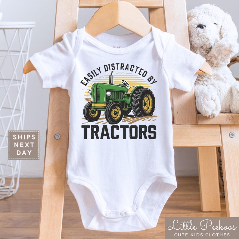 K&ouml;nnte beinhalten: Wei&szlig;er Baby-Body mit einer gr&uuml;nen Traktor-Grafik und dem Text "EASILY DISTRACTED BY TRACTORS". Der Traktor hat gelbe R&auml;der und ist vor einem gelb-orangen Hintergrund platziert. Der Body h&auml;ngt an einem Holzb&uuml;gel.