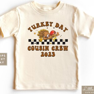 May include: A beige t-shirt with a brown turkey graphic and the text "Turkey Day Cousin Crew 2023".