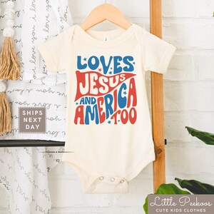 May include: Cream-colored baby onesie with the text "LOVES JESUS AND AMERICA TOO" in red and blue. The onesie is hanging on a wooden hanger. The text is in a retro font with star accents.