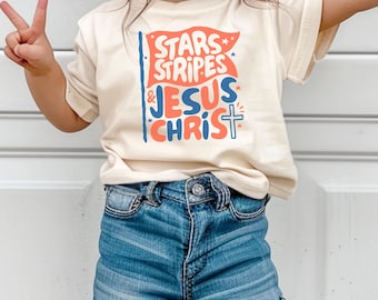 Puede incluir: Una camiseta color crema con el texto "Stars, Stripes & Jesus Christ" en rojo, azul y blanco. La camiseta se combina con pantalones cortos de mezclilla. La imagen muestra a un niño usando la camiseta y haciendo un signo de paz.