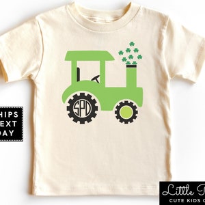 May include: A green tractor with a black gear design on the wheels and a black monogram "SPD" on the front wheel. The tractor has a green cab and a black chimney with green shamrocks coming out of it. The tractor is on a cream colored t-shirt.