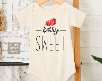 May include: Cream-colored baby onesie with the words "berry SWEET" in black script and a watercolor illustration of two red strawberries. The onesie is hanging on a wooden hanger.