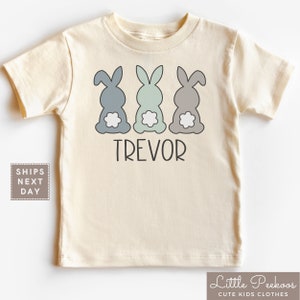 May include: A cream colored short-sleeved t-shirt with three gray, blue, and green bunny rabbits with white tails. The text "TREVOR" is printed below the bunnies.