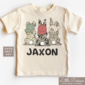 May include: A beige short-sleeved t-shirt with a camouflage pattern and three bunnies wearing hats. The bunnies are wearing a green, red, and blue hat. The text "Jaxon" is printed on the shirt.