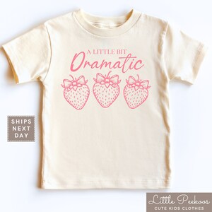 May include: A light beige t-shirt with a pink graphic that says "A Little Bit Dramatic" with three strawberries with bows underneath the text.
