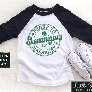 May include: White and black baseball tee shirt with green shamrock design. The shirt says "Prone to Shenanigans and Malarkey".