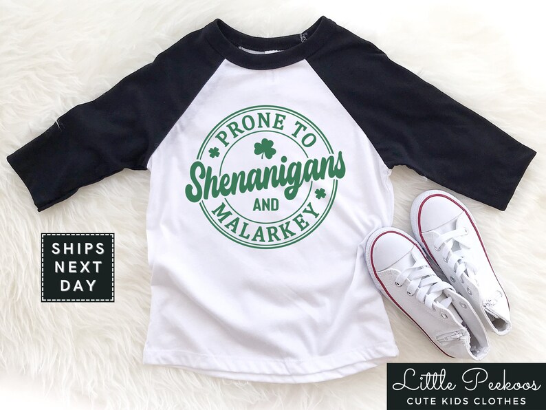 May include: White and black baseball tee shirt with green shamrock design. The shirt says "Prone to Shenanigans and Malarkey".