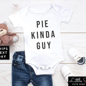 May include: White baby onesie with black text that reads "Pie Kinda Guy". The onesie is laying on a white wooden surface with a pair of blue jeans, a brown teddy bear, and two white baby shoes.