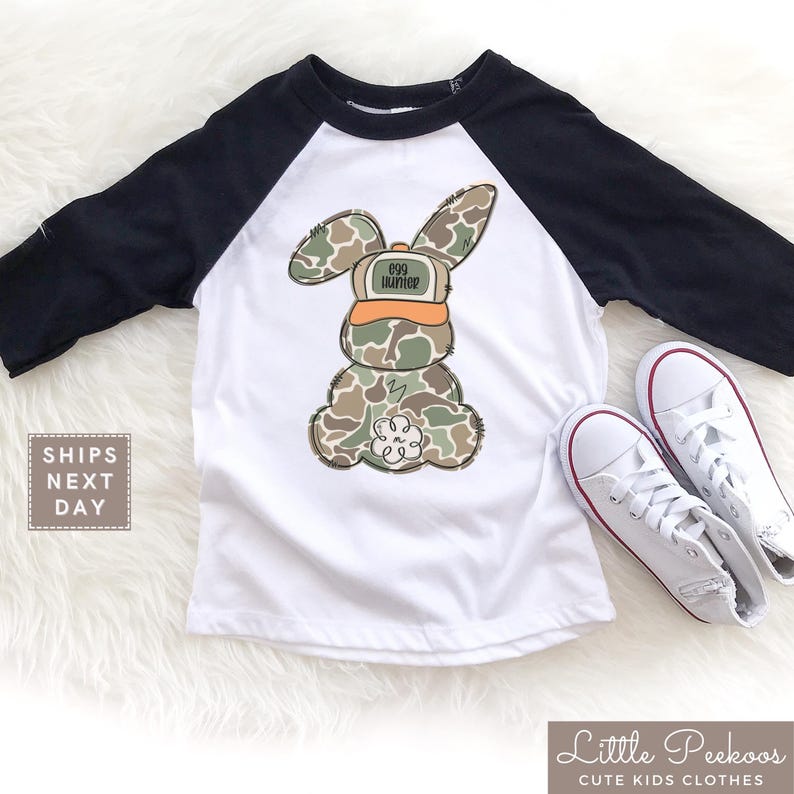May include: A white and black baseball tee shirt with a camouflage bunny rabbit wearing a hat that says "Egg Hunter".