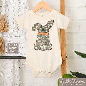 May include: A white baby onesie with a camouflage bunny rabbit wearing a hat that says "099 Hunter".
