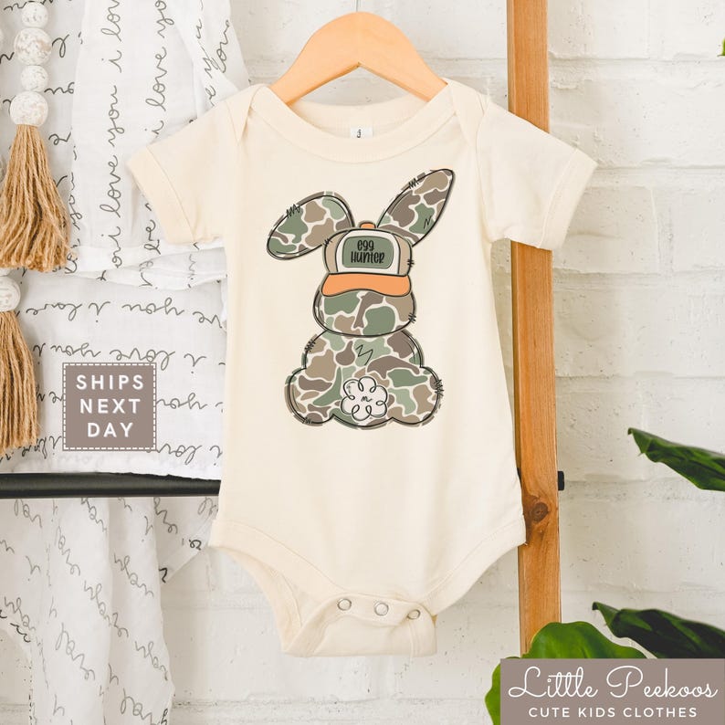 May include: A white baby onesie with a camouflage bunny rabbit wearing a hat that says "099 Hunter".