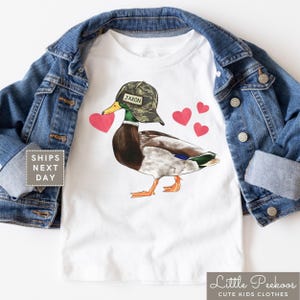 May include: White t-shirt with a cartoon duck wearing a camouflage hat with the name Jaxon on it. The duck is holding a red heart in its beak and there are several red hearts around it.