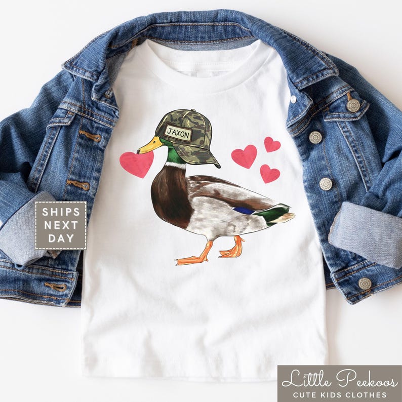 May include: White t-shirt with a cartoon duck wearing a camouflage hat with the name Jaxon on it. The duck is holding a red heart in its beak and there are several red hearts around it.