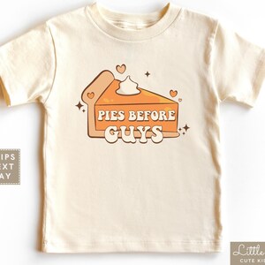 May include: A cream-colored short-sleeved t-shirt with a graphic of a slice of pumpkin pie with the text "Pies Before Guys" in orange and brown letters.