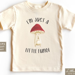 May include: A beige short-sleeved t-shirt with a cartoon mushroom graphic and the text "I'm just a little fungi".