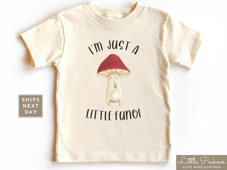 May include: A beige short-sleeved t-shirt with a cartoon mushroom graphic and the text "I'm just a little fungi".