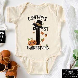 May include: A white onesie with a black graphic of a pilgrim hat on top of the number one. The graphic also includes the text "Cameron's 1st Thanksgiving 2022".