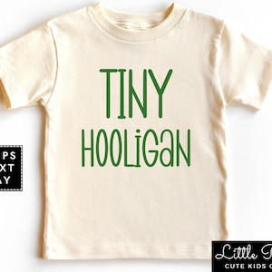 May include: A cream colored short-sleeved t-shirt with the text "TINY HOOLIGAN" in green lettering.