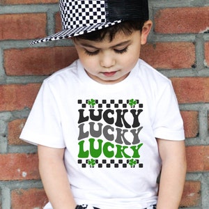 May include: A white t-shirt with a black and white checkered border and green shamrocks. The shirt has the word "LUCKY" written three times in a retro font, with each word in a different color: black, gray, and green.