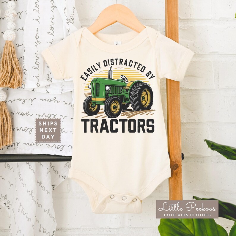 K&ouml;nnte beinhalten: Cremefarbener Baby-Body mit einer gr&uuml;nen Traktor-Grafik und dem Text "EASILY DISTRACTED BY TRACTORS". Der Traktor hat gelbe R&auml;der und ist vor einem gelb-orange gestreiften Hintergrund platziert. Der Body h&auml;ngt an einem Holzb&uuml;gel.
