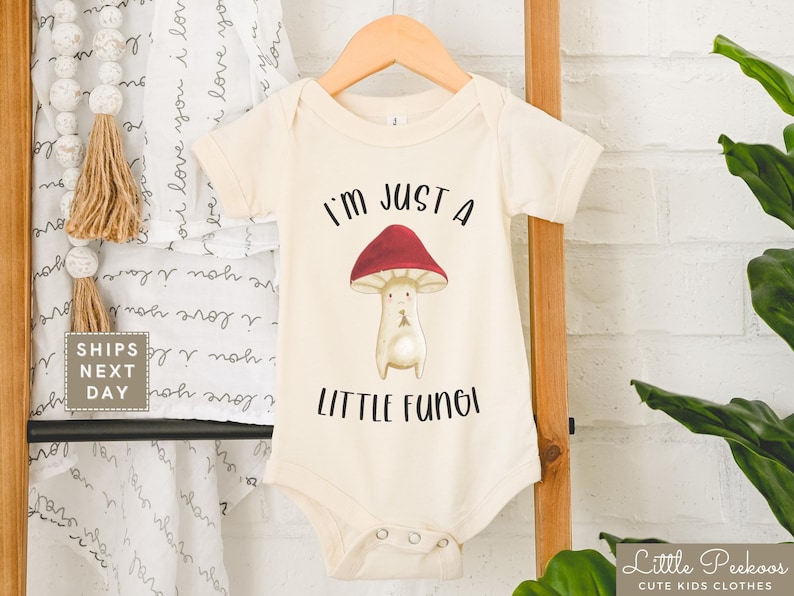 May include: A cream-colored baby onesie with short sleeves features a cartoon mushroom with a red cap and the text "I'm just a little fungi".