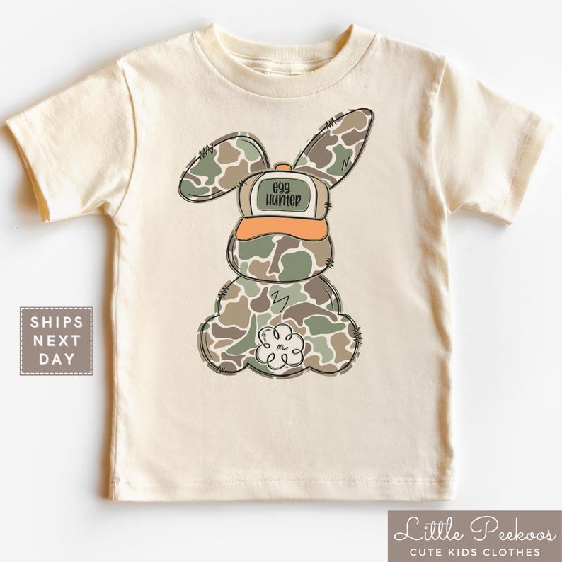 May include: A beige short-sleeved t-shirt with a cartoon bunny wearing a camouflage pattern and a hat that says "egg hunter".