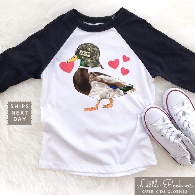 May include: A white and black baseball tee shirt with a cartoon duck wearing a camouflage hat that says "Jaxon". The duck is holding a pink heart in its beak and there are pink hearts scattered around it.