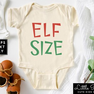 May include: A white baby onesie with the words "ELF SIZE" in red and green letters. The onesie is on a white background with a white blanket and brown shoes.
