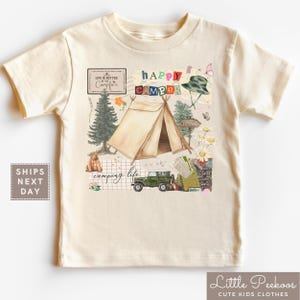 May include: Cream-colored t-shirt featuring a camping-themed graphic. The design includes a tent, a green jeep, a bear, and the words "Happy Camper" and "Camping Life." The shirt is made of soft material.
