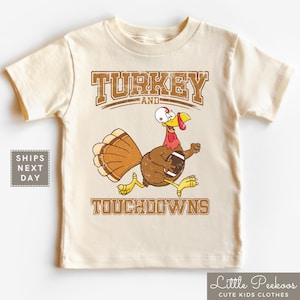 May include: A beige short-sleeved t-shirt with a cartoon turkey holding a football. The shirt has the text "TURKEY AND TOUCHDOWNS" in a vintage style font.