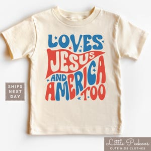 May include: Cream-colored t-shirt with the words "LOVES JESUS AND AMERICA TOO" in red and blue, with star accents. The shirt is a children's clothing item.