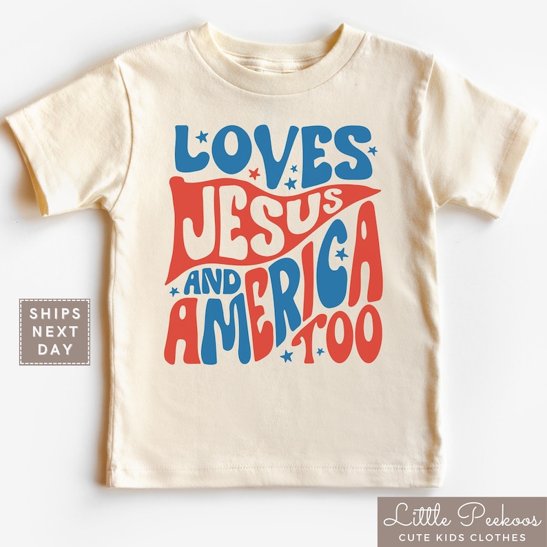 May include: Cream-colored t-shirt with the words "LOVES JESUS AND AMERICA TOO" in red and blue, with star accents. The shirt is a children's clothing item.
