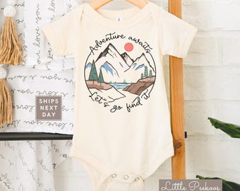 May include: Cream-colored baby onesie with a mountain and sun graphic. The design includes the text "Adventure awaits" and "Let's go find it" in a circular design. The onesie is hanging on a wooden hanger.