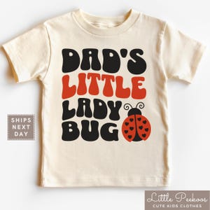 May include: Cream-colored toddler t-shirt with the text "DAD'S LITTLE LADY BUG" in black and red, featuring a ladybug graphic. The shirt also has the text "Little Peekoos Cute Kids Clothes".