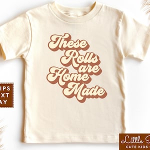 May include: A cream colored t-shirt with a retro-style graphic that says "These Rolls Are Home Made" in brown and cream colors.