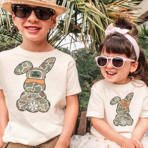 May include: Two children wearing cream colored t-shirts with camouflage bunny designs. The boy's shirt says "egg hunter" on the bunny's hat. The girl's bunny has a bow on its ear. Both children are wearing sunglasses.