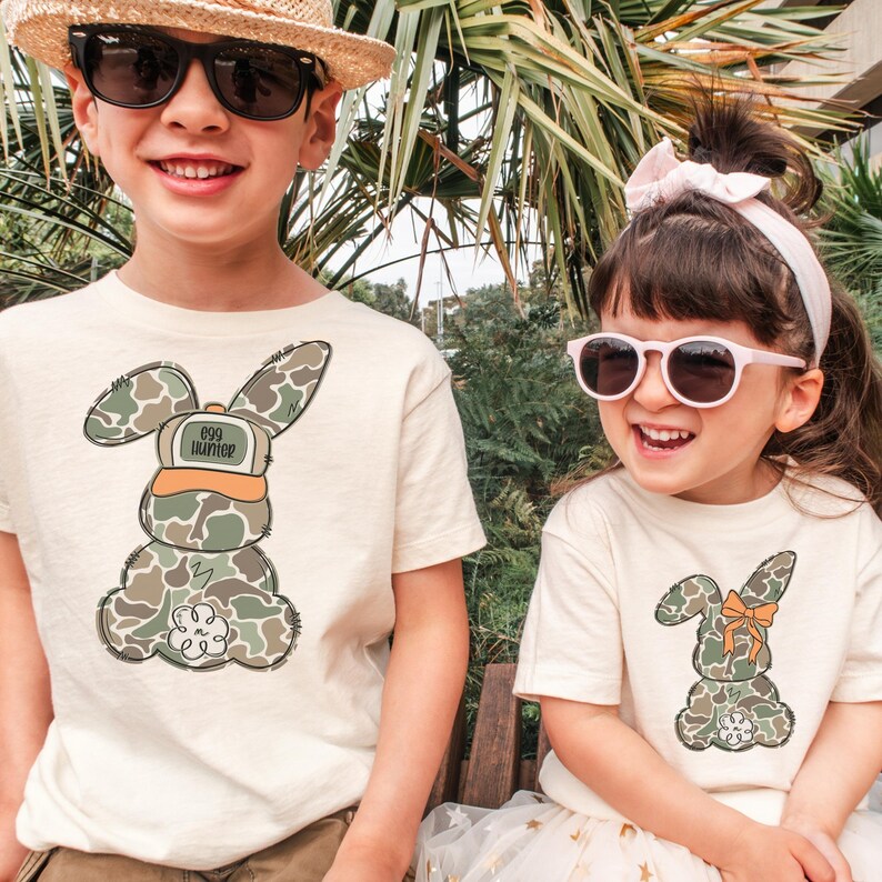 May include: Two children wearing cream colored t-shirts with camouflage bunny designs. The boy's shirt says "egg hunter" on the bunny's hat. The girl's bunny has a bow on its ear. Both children are wearing sunglasses.