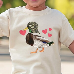 May include: A white t-shirt with a cartoon image of a duck wearing a camouflage hat with the name "Jaxon" on it. The duck is holding a pink heart in its beak and there are three pink hearts around it.